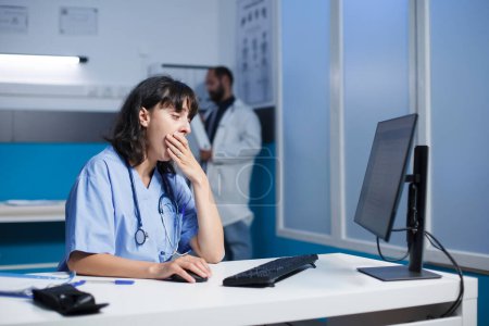 Fatigued female practitioner in modern hospital office, using digital technology for collaboration and communication. Dedicated nurse in blue scrubs tiredly reviewing medical data on desktop computer.