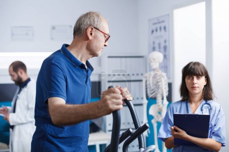 Female nurse assists senior patient with receiving alternative therapy. Medical specialist with clipboard looks at camera while elderly man exercises on stationary bicycle for physical recovery.