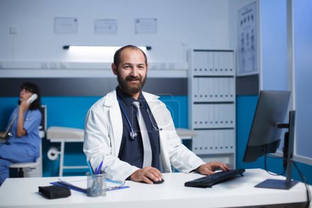 Photo for Middle aged doctor smiling at his desk in a medical office. Caucasian male physician appears to be wearing a white lab coat, female nurse is in the background on a call. Portrait shot. - Royalty Free Image