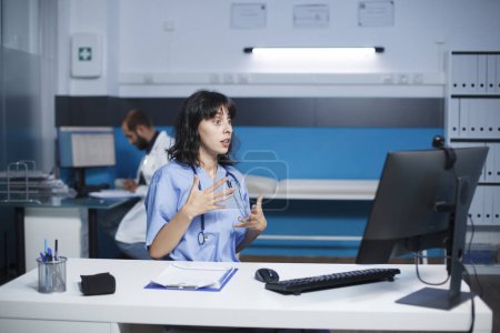 Téléchargez les photos : Infirmière dans un hôpital communique par un appel vidéo à l'aide de son ordinateur de bureau. Femme caucasienne assise dans le bureau de la clinique ayant une réunion virtuelle. Collaboration et technologie dans le domaine de la santé. - en image libre de droit