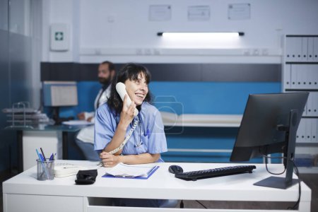 Foto de Una practicante sonriente sentada en el escritorio de la clínica está hablando por teléfono fijo. Enfermera caucásica en llamada telefónica mientras usa una computadora de escritorio en una habitación de hospital. - Imagen libre de derechos