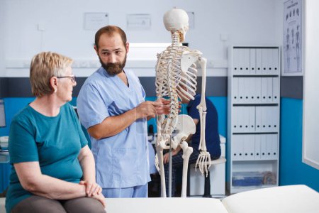 Nurse demonstrates spinal cord on human skeleton to elderly woman in physiotherapy clinic. Male doctor discusses anatomy and spinal bones with retired old patient seated on hospital bed.