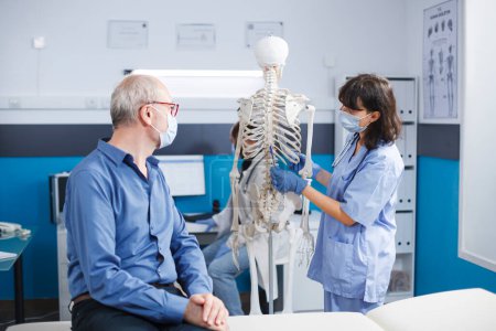 Nurse wearing face mask is describing bones in human skeleton to elderly guy in cabinet. They are also discussing osteopathy anatomy and spinal cord discomfort. Doctor doing a physical assessment.