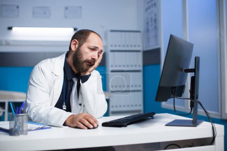Bärtiger kaukasischer Mann im Laborkittel, der müde medizinische Informationen auf seinem Desktop-PC durchsieht. Männlicher Arzt sieht abgedroschen aus und benutzt Computer in einem Krankenhauszimmer.