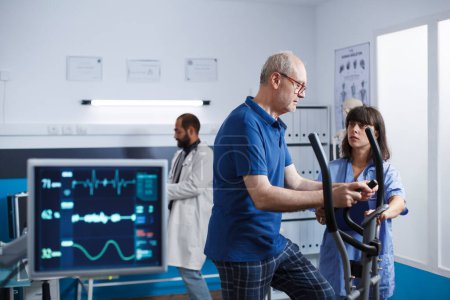 Nurse practitioner giving assistance to old man using stationary bike for physical activity and recovery. Medical assistant helping male patient with exercise bicycle for physiotherapy.