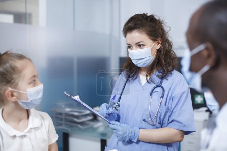 Téléchargez les photos : Au cabinet médical, une femme médecin portant des blouses bleues s'entretient avec une jeune patiente. Les deux individus portent des masques faciaux. Le matériel médical, y compris un stéthoscope, est visible. - en image libre de droit
