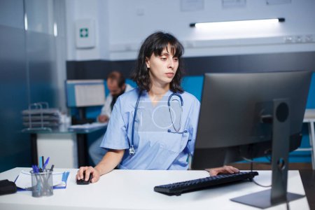 Detailliertes Bild einer kaukasischen Krankenschwester, die in einem Krankenhausbüro einen Desktop-Computer bedient. Eine Ärztin mit blauem Peeling überprüft medizinische Akten auf einem Computer in einem Klinikzimmer.