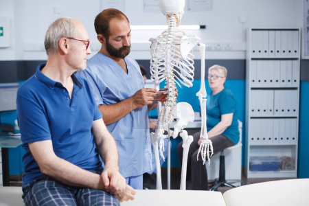 In physiotherapy clinic, orthopedist explains spinal cord on human skeleton to elderly patient. Doctor discussing anatomy and spinal bones with old man in preparation for osteopathy diagnostic.