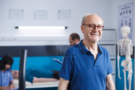 Photo for Elderly man with glasses standing in a physiotherapy facility. Retired senior patient smiling at camera as he gets ready for his recovery at clinic for rehabilitation treatment through fitness. - Royalty Free Image