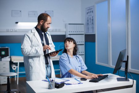 Kaukasische Ärztin und Gesundheitshelferin arbeiten in einer Klinik zusammen. Sie diskutieren digitale medizinische Bilder auf Tablet und Desktop-Computer. Medizinische Geräte sind sichtbar.