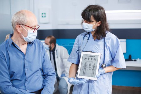Healthcare specialist describes human skeleton image on tablet to senior patient with face mask at medical exam. Female physician grasps device with osteopathy diagnosis, showing bones examination.