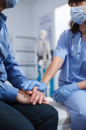 Close-up of female nurse holding hands of senior patient at hospital office. Medical practitioner wearing gloves assists retired old man after a chiropractic examination and diagnosis.