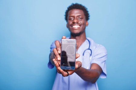 Photo for Male nurse looking at camera while carrying cell phone with blank screen, against blue background. Demonstrating medical expertise, black man wears scrubs and stethoscope while holding mobile device. - Royalty Free Image