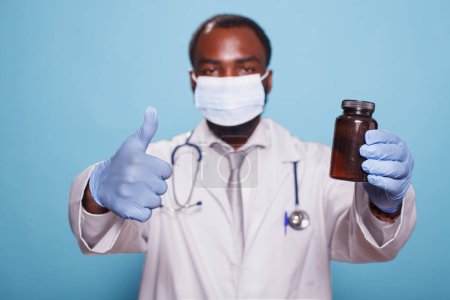 Photo for Portrait of medical specialist in a face mask, showing thumbs up while gripping pills bottle. Black doctor with stethoscope and gloves approving prescription painkillers with hand gestures. - Royalty Free Image