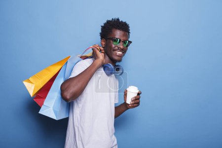 Téléchargez les photos : Joyeux Afro-Américain tenant une tasse de bonheur après une virée shopping. Un homme optimiste portant des lunettes de soleil et des écouteurs, tenant des sacs avec des produits du Black Friday. - en image libre de droit