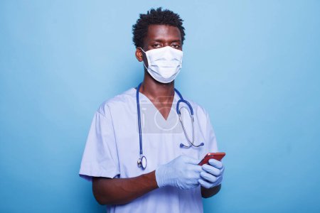 Photo for Close-up of nurse wearing face mask, holding smartphone for healthcare information during pandemic. African American medical professional stands confidently against blue background with mobile device. - Royalty Free Image