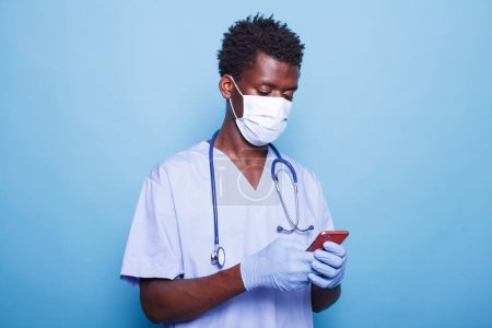 Photo for African american medical assistant in blue scrubs grasping smartphone in hand. Male healthcare specialist wearing face mask and gloves for covid 19 protection while having a mobile device. - Royalty Free Image