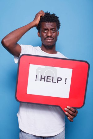 Photo for Bewildered african american individual holding voice bubble with text message asking for help. Portrait of black guy with hand on his head is grasping a red cardboard sign. - Royalty Free Image
