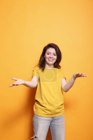Photo for Joyful young woman with expressive positive attitude welcoming with raised arms in studio. Portrait of brunette lady smiling, being friendly and sociable, doing a greeting gesture. - Royalty Free Image