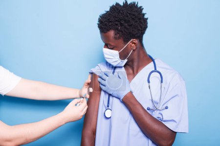 Photo for Caucasian person administering vaccine injection to African American nurse against blue background. Close-up of a doctor giving vaccination to black man, ensuring healthcare and covid 19 prevention. - Royalty Free Image