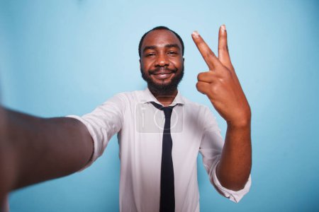 Téléchargez les photos : POV de jeune entrepreneur masculin faisant vlog en ligne pour la publicité sur les médias sociaux. Homme d'affaires noir prenant un selfie avec signe de paix geste de la main devant fond bleu isolé. - en image libre de droit