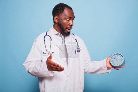 Médico general afroamericano escandalosamente mirando el reloj antes de una consulta médica del paciente. Médico negro parece estar en pánico, corriendo y experimentando una emergencia de salud.