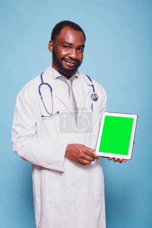 Foto de Retrato del hombre negro con un estetoscopio y una bata de laboratorio sostiene una tableta con pantalla verde. Dispositivo de agarre médico afroamericano que muestra una plantilla de maqueta cromakey en blanco. - Imagen libre de derechos