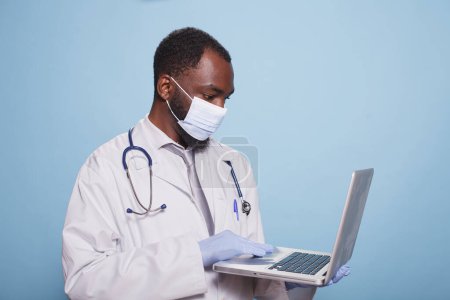 Téléchargez les photos : Médecin avec gants en latex et masque facial tenant un ordinateur portable pour examiner les résultats d'un diagnostic clinique. L'ordinateur portable est tenu par un médecin qui porte une blouse et un stéthoscope.. - en image libre de droit