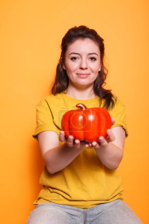 Téléchargez les photos : Portrait de femme caucasienne tenant un poivron et regardant dans la caméra debout en studio avec un fond orange. Jeune femme ayant une alimentation saine, manger des légumes. Concept végétarien biologique. - en image libre de droit