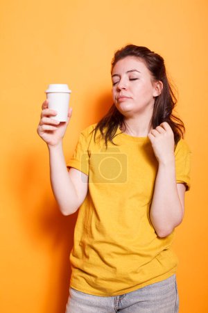 Müde Frau posiert vor orangefarbenem Hintergrund in lässiger Kleidung, hält Kaffeetasse in der Hand, entspannt mit geschlossenen Augen. Schläfriger Ausdruck einer jungen brünetten Dame zeigt Erschöpfung und Müdigkeit.