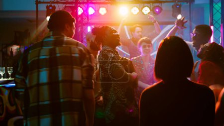 Téléchargez les photos : Des amis heureux dansant sur une musique funky, faisant la fête avec DJ sur scène et projecteurs. Groupe de personnes diverses sautant sur la piste de danse et s'amusant à l'événement de clubbing. Coup de main. - en image libre de droit