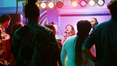 Foto de Mujer feliz haciendo karaoke en la fiesta, cantando canciones con DJ delante de amigos en la pista de baile. Chica segura de divertirse con perfromance espectáculo en vivo junto a la estación de mezcla electrónica en la discoteca. - Imagen libre de derechos