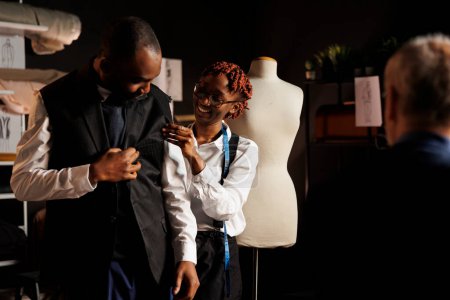 Photo for African american seamstress helping client try on sartorial piece after finishing needlework. Customer happy with bespoken elegant attire design in atelier shop run by senior tailor - Royalty Free Image