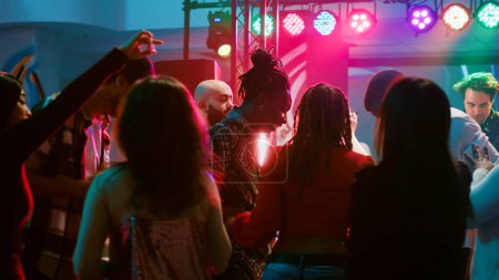 Young adults doing cool dance moves at club, partying together and having fun on electronic music. Diverse crowd of people enjoying night out with live performance on dance floor.