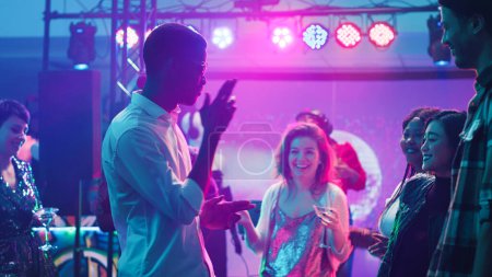 Foto de Hombre casual mostrando movimientos de baile funky en el club, moviéndose en la música electrónica y divirtiéndose con amigos. Grupo de personas modernas de fiesta con DJ en la estación de audio, lleno de pista de baile. Trípode tiro. - Imagen libre de derechos
