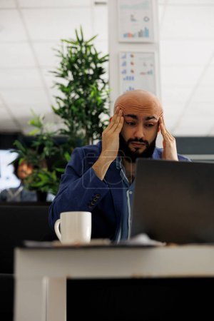 Exhausted arab entrepreneur with migraine writing business plan on laptop in office. Overworked employee having headache while analyzing start up company marketing strategy