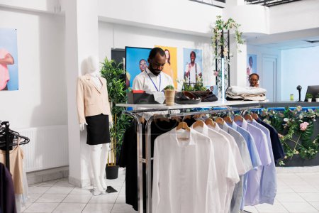 Téléchargez les photos : Shopping centre commercial assistant afro-américain travaillant dans un magasin de mode, la mise en vente de vêtements. Showroom homme employé vérifier la marchandise et afficher des vêtements sur les cintres pour les clients - en image libre de droit