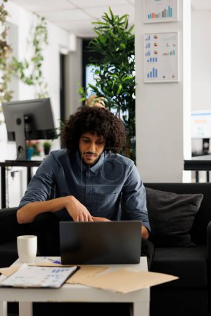 Photo for Arab executive manager working at start up project development in business office. Young businessman sitting on couch at workspace and checking marketing research on laptop - Royalty Free Image