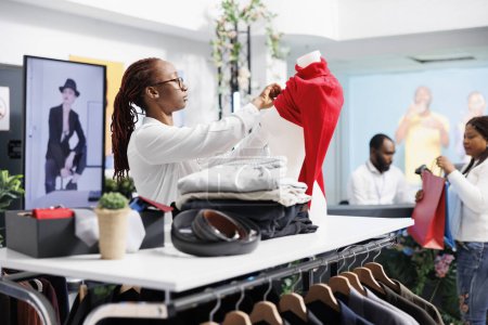 Téléchargez les photos : Mannequin de femme afro-américaine en pull rouge dans un magasin de vêtements. Assistante de centre commercial mettant une blouse décontractée féminine sur un mannequin pour afficher des vêtements à vendre - en image libre de droit