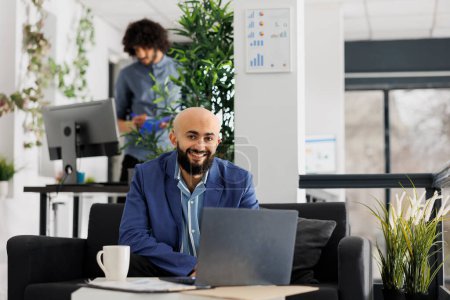 Téléchargez les photos : Entrepreneur souriant travaillant sur un ordinateur portable et regardant la caméra dans le bureau de l'entreprise en démarrage. Gestionnaire de projet réussi en utilisant un ordinateur portable assis sur le canapé portrait - en image libre de droit