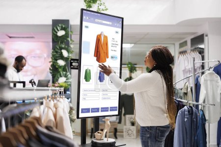 Käuferin wählt Kleidung an Bord aus, mittels interaktivem Touchscreen-Monitor im Bekleidungsgeschäft. Junge Boutiquekunden kaufen Modeartikel im Selbstbestellservice, im Einzelhandel.