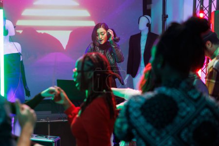 Foto de Mujer dj actuando en el escenario durante la fiesta disco en discoteca. Músico asiático sosteniendo auriculares y mezclando música electrónica mientras la multitud festeja y baila en la pista de baile en el club - Imagen libre de derechos