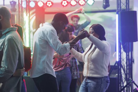 Photo for Young man and woman friends holding hands while dancing at nightclub party. African american couple clubbing and moving on dancefloor while attending discotheque gathering event - Royalty Free Image