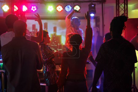 Téléchargez les photos : Clubbers faire la fête et danser tout en assistant à la performance dj dans la boîte de nuit. La foule des jeunes debout les mains levées sur le dancefloor illuminé de projecteurs à la discothèque - en image libre de droit