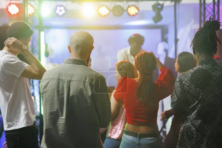 Téléchargez les photos : Jeunes debout sur la piste de danse tout en assistant à un concert de musique électronique dans une boîte de nuit. Divers hommes et femmes foule dansant tandis que dj se produisant sur scène avec des projecteurs colorés - en image libre de droit