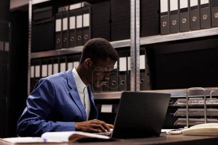 Téléchargez les photos : Homme d'affaires afro-américain analysant des dossiers administratifs, lisant un rapport comptable dans un dépôt d'entreprise. Comptable travaillant tard dans la nuit à la bureaucratie record dans la salle de stockage - en image libre de droit