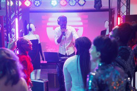Photo for Young man dj mixing music using controller and singing in microphone on club stage with spotlights. African american musician entertaining clubbers crowd at party in nightclub - Royalty Free Image
