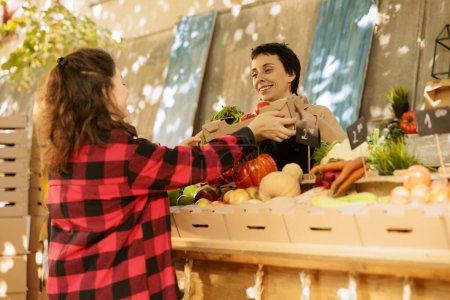 Foto de Joven compradora sosteniendo caja de cartón con frutas y verduras orgánicas cultivadas localmente. Vendedor sonriente con delantal que vende verduras naturales saludables en el mercado local de agricultores. - Imagen libre de derechos