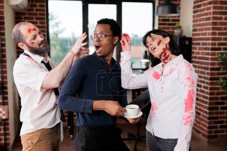 Employees wearing zombie costumes goofing around with manager in office, pretending to possess him. Team leader and coworkers dressed as undead creatures having fun during Halloween event at work