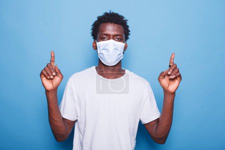 Photo for Portrait of black man wearing mask for coronavirus protection is pointing up with his two index fingers. African american guy wearing face mask while doing sign with hands in the studio. - Royalty Free Image